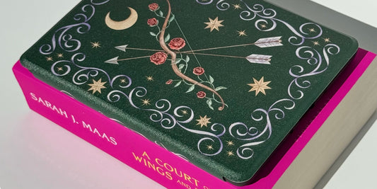 The Huntress Kindle case, gold moon and roses on deep green, beside a Sarah J. Maas paperback.